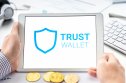 Trust Wallet Token A tablet showing the Trust Wallet logo next to a wallet full of physical cryptocurrencies