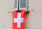 The Swiss franc is on the move again but the SNB has a massive balancing act to handle for this export-intensive currency. Red & white Swiss flag draped under window