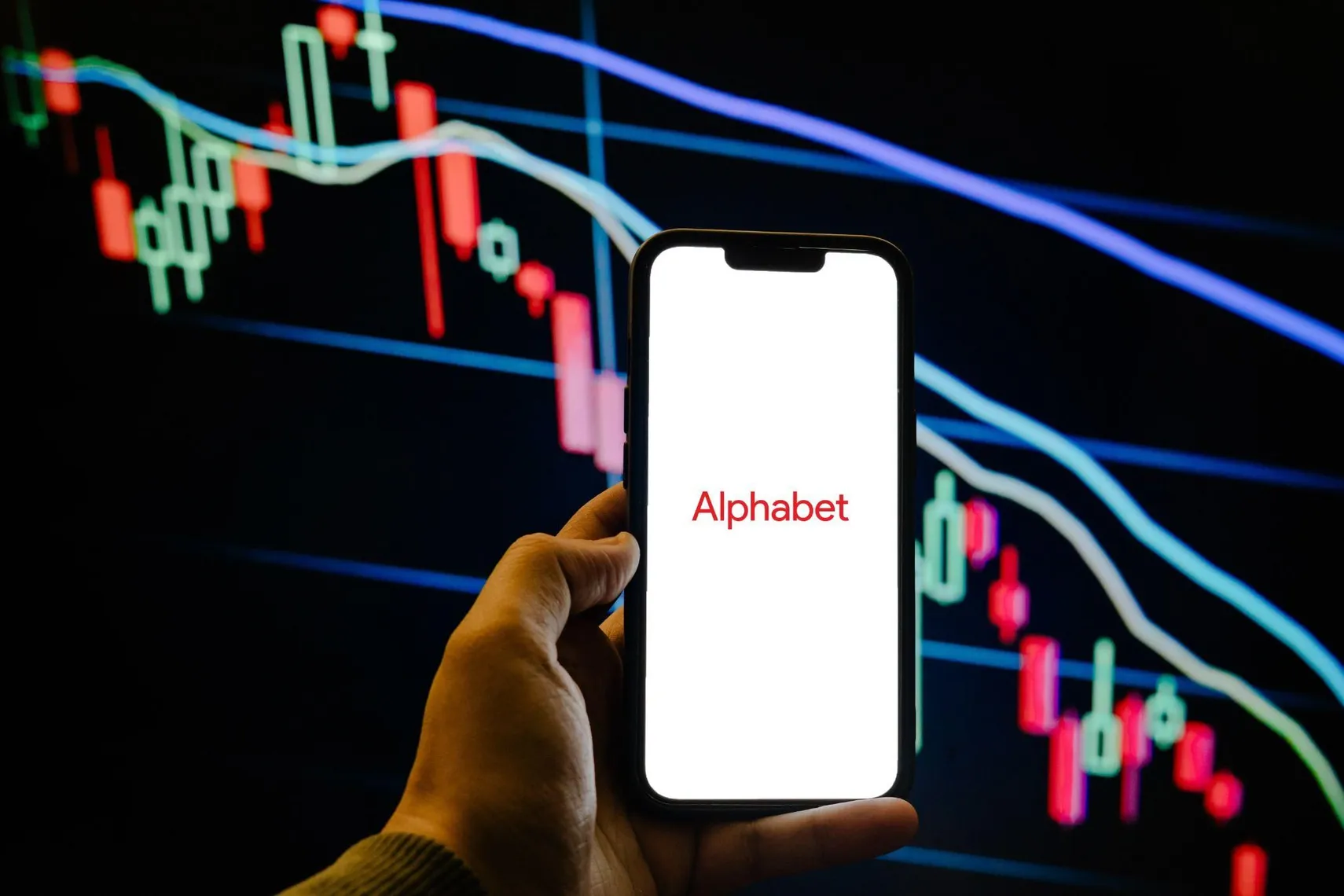 Alphabet logo on smartphone with stock market chart Smartphone displaying the Alphabet logo in front of a bearish candlestick stock chart, representing the technology company and its stock market performance.