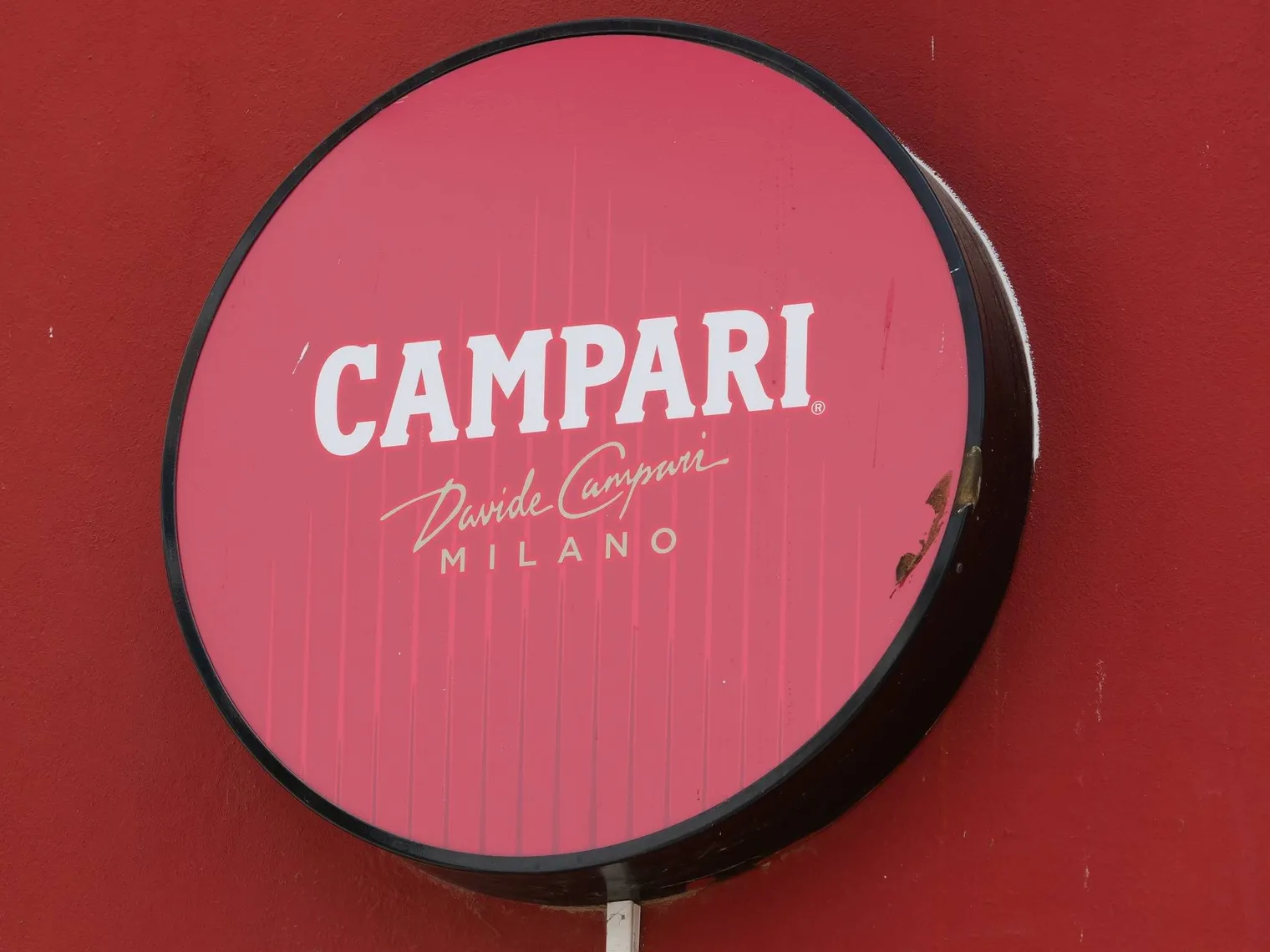Round illuminated Campari sign mounted on a red wall with white Campari Milano branding Round illuminated Campari sign mounted on a red wall with white Campari Milano branding