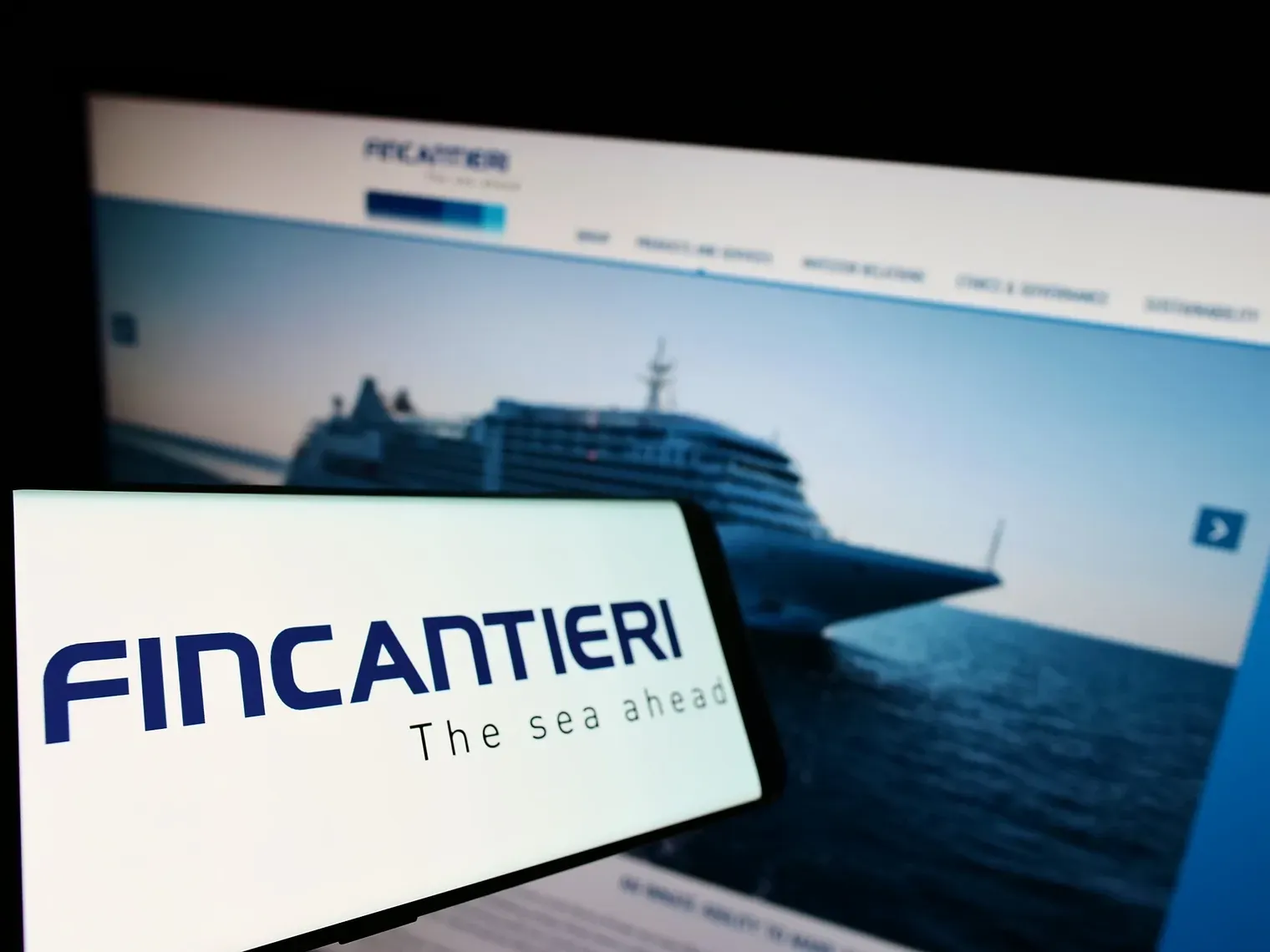 Smartphone che mostra il logo Fincantieri con un'immagine sfocata di una nave da crociera su un sito web sullo sfondo