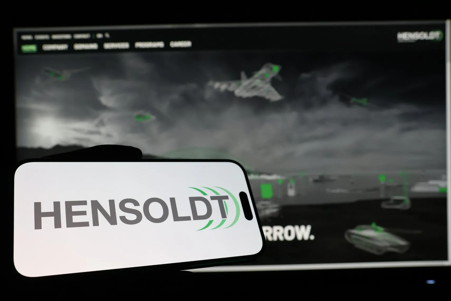 HENSOLDT logo on a mobile device with military technology visuals, representing the defense and security industry HENSOLDT logo on a mobile device with military technology visuals, representing the defense and security industry