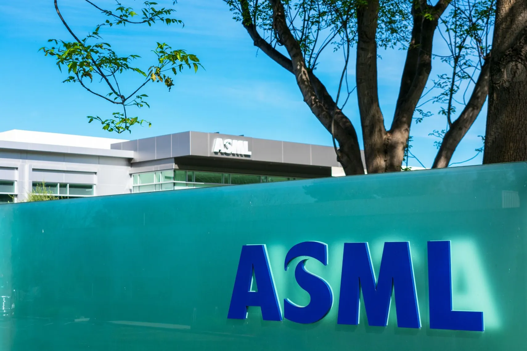ASML office building with the company logo displayed on a turquoise panel in front of a modern facility, surrounded by trees under a clear blue sky. ASML office building with the company logo displayed on a turquoise panel in front of a modern facility, surrounded by trees under a clear blue sky.