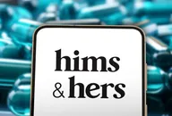 Smartphone displaying the Hims & Hers logo in front of blurred prescription capsules, representing the digital healthcare and telemedicine company.