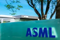 ASML office building with the company logo displayed on a turquoise panel in front of a modern facility, surrounded by trees under a clear blue sky.