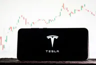 Image of Tesla logo on a smartphone against a background of stock market trends
