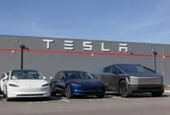 Tesla vehicles parked in front of a Tesla facility building with the company logo displayed on the wall under a clear blue sky