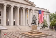 US Treasury Department building in Washington, DC, with its neoclassical columns, a large American flag on the facade and the Albert Gallatin statue in the forecourt