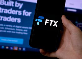 A smartphone displays the logo of FTX in front of a screen showing the FTX website