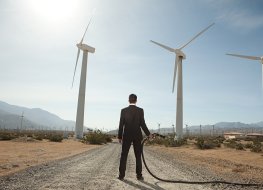 A businessman at a wind farm with a gasoline pump in his hand.