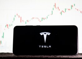 Image of Tesla logo on a smartphone against a background of stock market trends