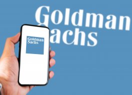 Hand holding a smart phone with Goldman Sachs logo on screen and blurry Goldman Sachs logo on the background.