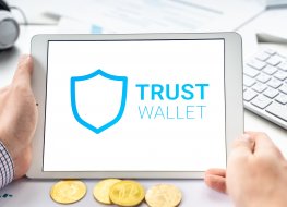 A tablet showing the Trust Wallet logo next to a wallet full of physical cryptocurrencies