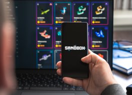 A photo of a hand holding a phone with Sandbox (SAND) logo.