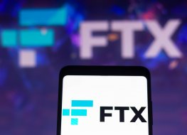 The FTX logo on a smartphone, which is in front of a computer screen also displaying the FTX name and logo