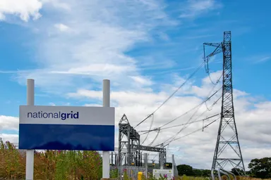 National Grid sign beside an electricity substation with transmission lines and pylons under a blue sky, representing UK energy infrastructure and power distribution. 