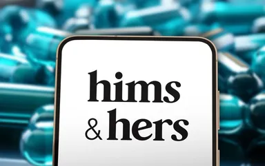 Smartphone displaying the Hims & Hers logo in front of blurred prescription capsules, representing the digital healthcare and telemedicine company.