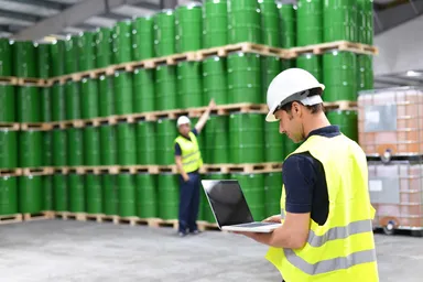 Warehouse workers wearing safety helmets and high-visibility vests inspecting stacked green industrial barrels