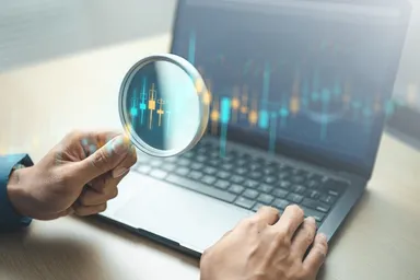 Hand holding a magnifying glass over a laptop displaying a candlestick trading chart, representing technical analysis and market research.
