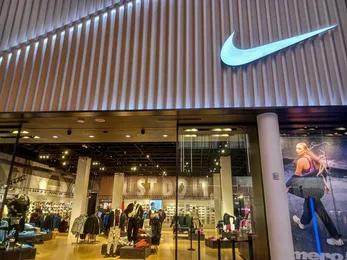 Nike retail store exterior with illuminated Nike logo above the entrance and sports apparel displayed inside.