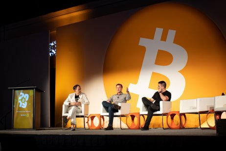 three people on stage with Bitcoin logo in the background