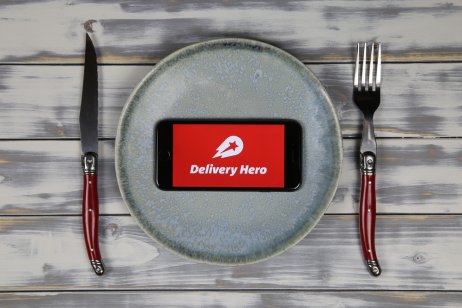 Close up of mobile phone screen with logo lettering of food delivery service hero on wood table with dish and cutlery