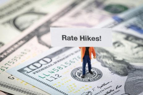 miniature man holding Rate Hikes sign standing on US Federal Reserve emblem on dollars banknote.