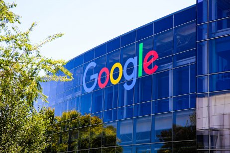 An exterior shot of the Googleplex building in Mountain View, California. it has a blue glass front with the multicoloured Google logo on it