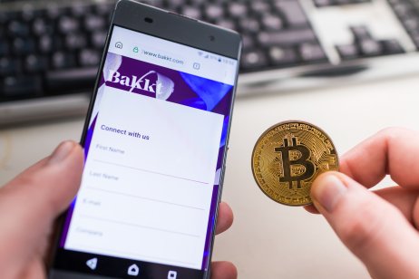 A customer holds a smartphone with a Bakkt Holdings logo in one hand a bitcoin token in the other