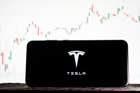 Image of Tesla logo on a smartphone against a background of stock market trends
