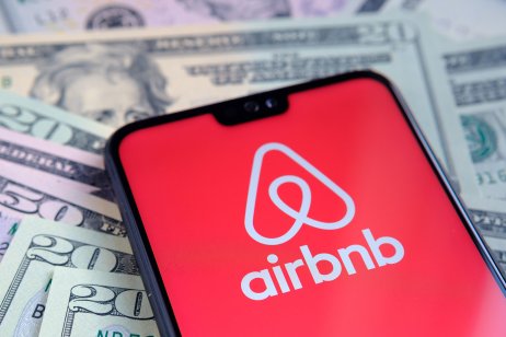 Airbnb stock price prediction: Is now the time to buy? Airbnb app logo seen on the screen of smartphone, placed on dollar bills. Concept for IPO and company profit.