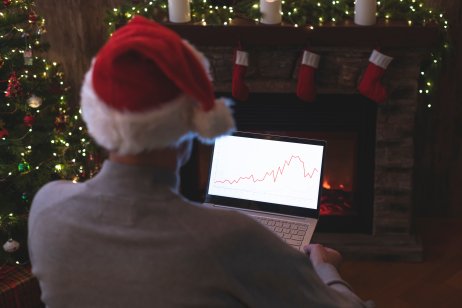 Man freelancer in santa claus hat working on laptop with graphs and charts on screen sitting near christmas tree and fireplace.