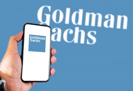 Hand holding a smart phone with Goldman Sachs logo on screen and blurry Goldman Sachs logo on the background.