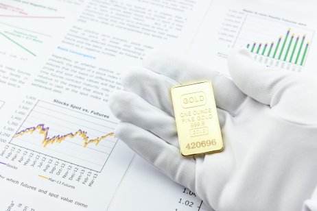 Hand holding one ounce of gold on top of financial market reports