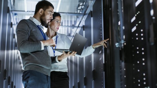 A male IT specialist holds a laptop and discusses work with a female server maintenance specialist. They are in the data center with the server rack open.