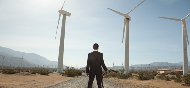A businessman at a wind farm with a gasoline pump in his hand.