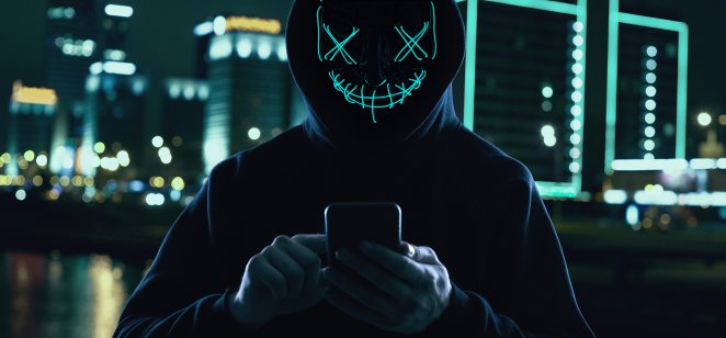 Anonymous in black hoodie and neon mask hacking into a smartphone. 