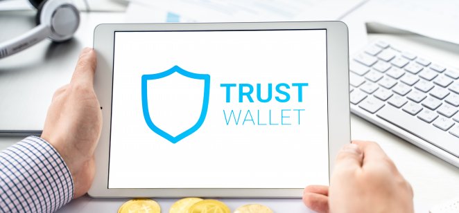 A tablet showing the Trust Wallet logo next to a wallet full of physical cryptocurrencies