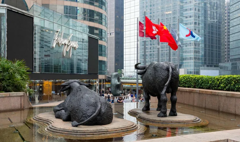 香港交易所外的牛雕塑與香港及中國國旗,背景為中環金融區辦公大樓,象徵香港股市與藍籌投資環境。 香港交易所外的牛雕塑與香港及中國國旗,背景為中環金融區辦公大樓,象徵香港股市與藍籌投資環境。