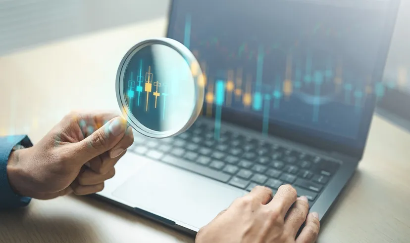 Hand holding a magnifying glass over a laptop displaying a candlestick trading chart, representing technical analysis and market research.
