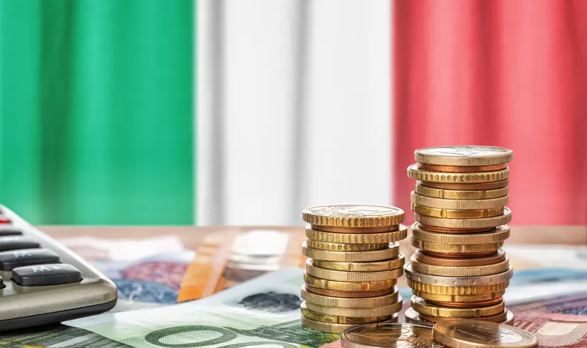 Pile di monete in euro su banconote con la bandiera italiana sullo sfondo.