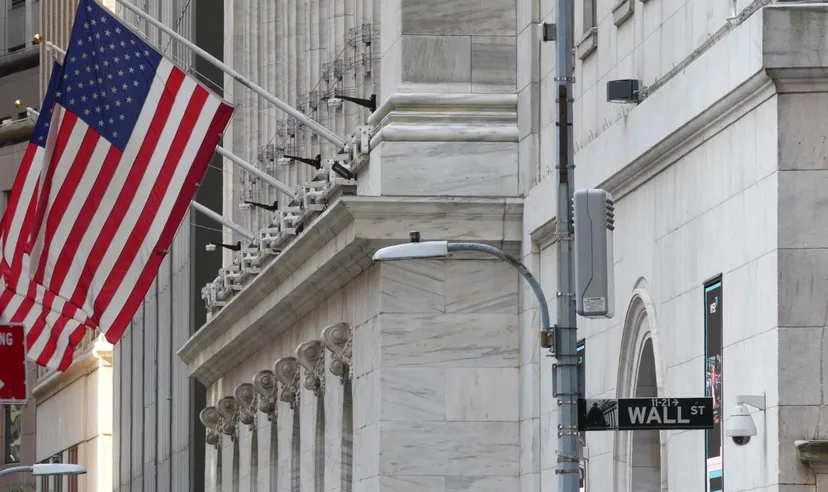 Wall Street sign beside the marble facade of the New York Stock Exchange building, with American flags flying from the exterior