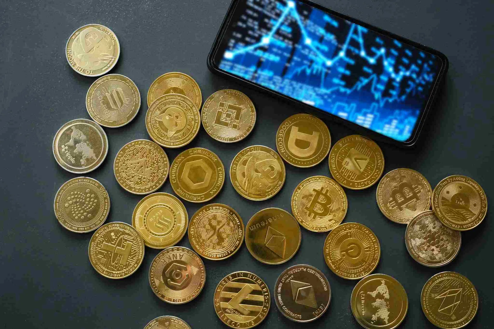 Collection of cryptocurrency coins beside a smartphone displaying a digital market chart, representing crypto trading, blockchain assets, and cryptocurrency markets.