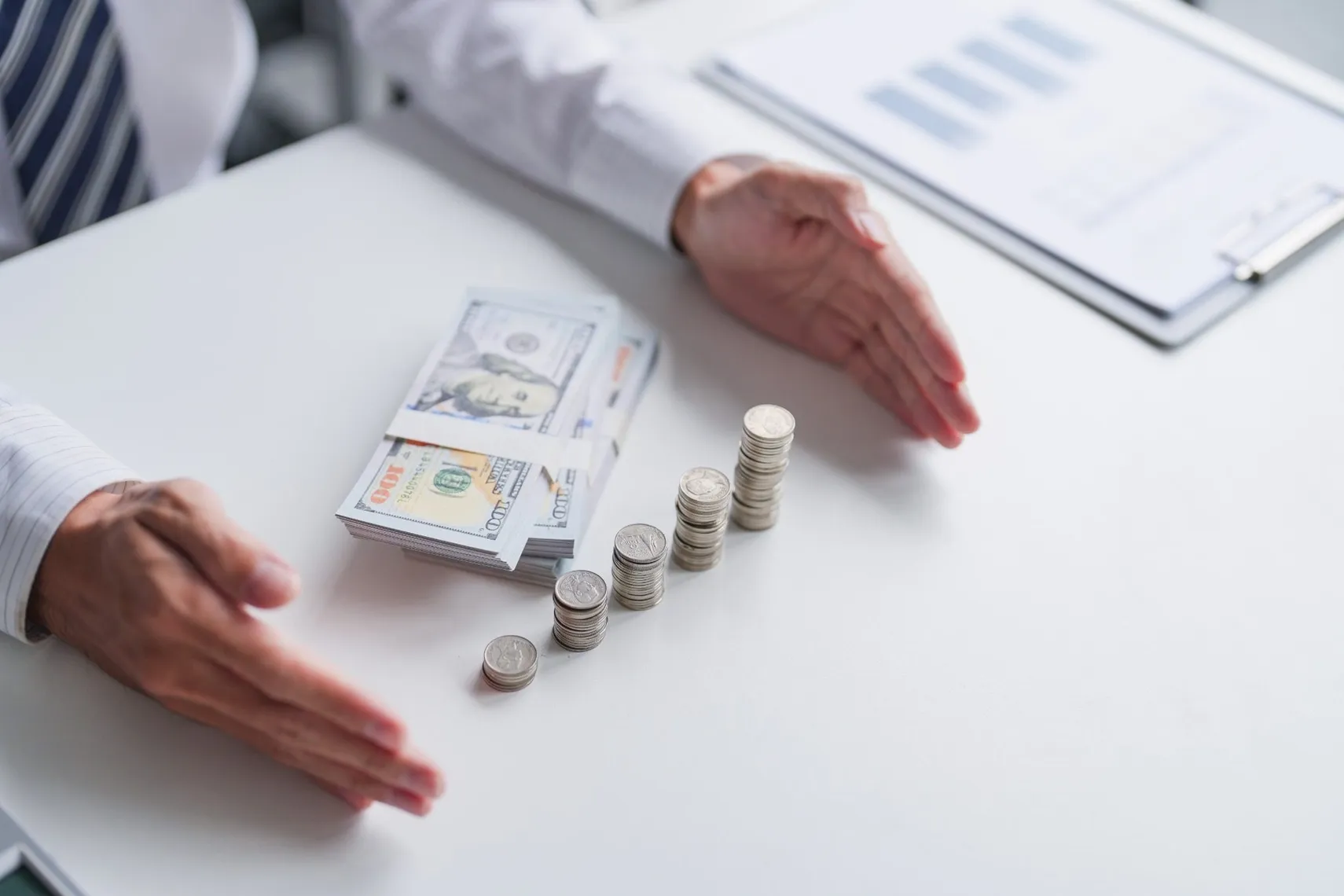 Cash and growing coin stacks on office desk (Source: Shutterstock) Hands framing stacks of coins and US dollar banknotes on an office desk, illustrating savings, investment growth, and personal finance planning.