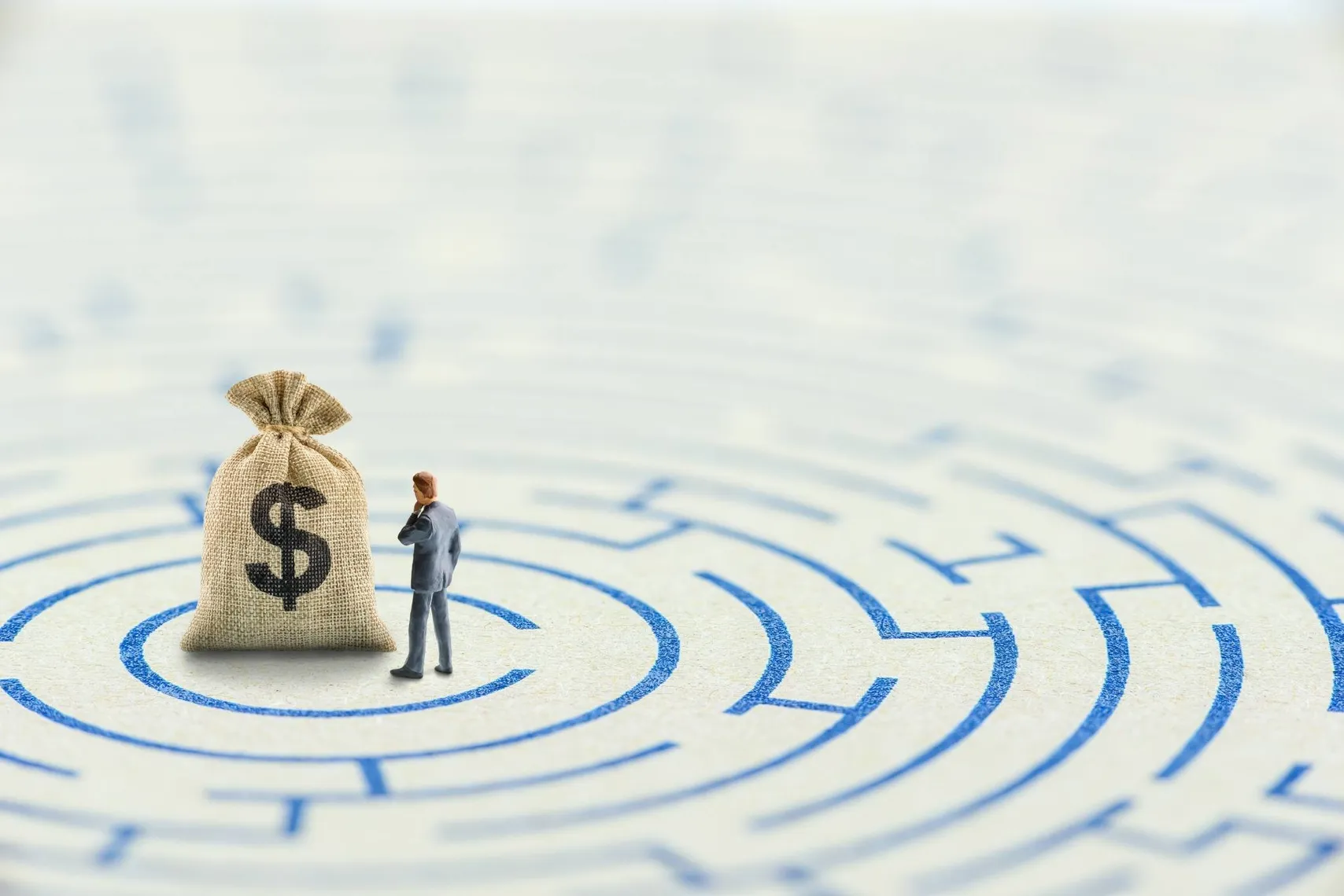 Money bag at the centre of a financial maze (Source: Shutterstock) Miniature person standing beside a money bag in the centre of a maze, symbolising financial planning, investment decisions, and wealth management.